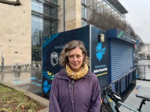 A woman wearing a purple coat stood in front of an 'Active Travel Hub' in Belfast. There is a bike beside her. 