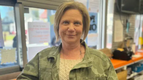 Nikki is wearing a green jacket and a beige jumper. She's leaning against a counter. She's smiling and has fair hair.