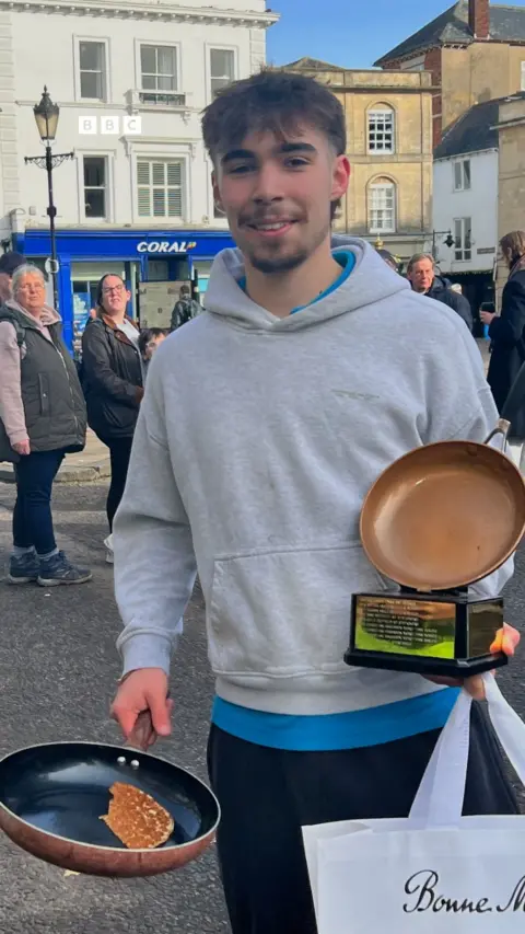 A man in a grey top holding a frying pan and a trophy.