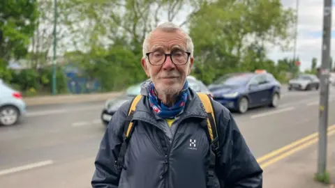 A portrait image of local Bernard Jarvis, 80, stood on the pavement in front of the road which crosses Lady Bay Bridge. There are cars in the background.