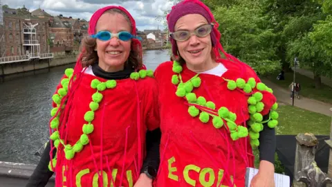 BBC/Olivia Richwald Two people standing side by side on a bridge, dressed as E.Coli. They are positioned in the foreground and are facing directly toward the camera. They are wearing bright, eye-catching red costumes which are decorated with green pom-pom-like shapes and long pink strands. Each person is also wearing swim goggles over their eyes, and both have distinctive head coverings in shades of pink or purple that match their outfits.