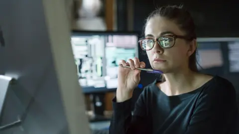 Woman wearing glasses and black top, holding a biro under chin and looking at a computer monitor, with other screens seen behind her