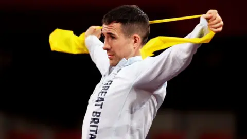 Getty Images A man in a white sports jacket stretches his arms wide behind his head with a yellow band between his hands. 