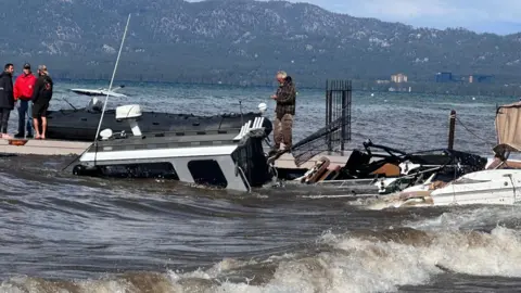 Several boats appear to have collided and capsized on Lake Tahoe. People are standing on top of the boats and one man appears to be on his cell phone. The mountains can be seen in the background.