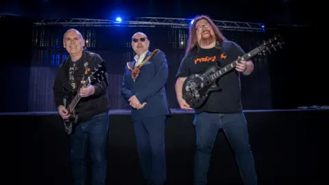 A man wearing a suit and mayoral chain of office is standing in between two other men who are both holding electric guitars. The two men with guitars are wearing jeans and black tops. Behind the three men is a lighting rig with blue lighting illuminating the scene.
