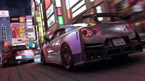 Playground Games Screenshot shows two sports cars speeding down a narrow urban road with tall buildings on either side. The fronts of the buildings have dozens of brightly lit signs advertising businesses within.