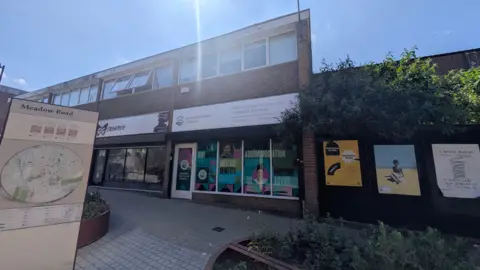 A two storey building with 4 units in a row. One unit belongs to Accommodation Concern, which has large colourful windows and a front door, all with pink, green and blue lettering. Words seen on the windows in large letters include accommodation, welfare benefits and housing advice. In the foreground there are established bushes and a public sign with a map of the local area titled Meadow Road, as well as a large paved area. 