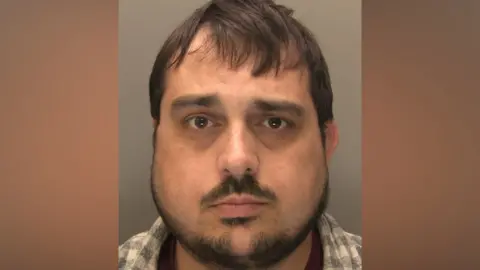 Police mug shot of a man with dark hair and a beard. He is wearing a checked shirt and looking directly at the camera.
