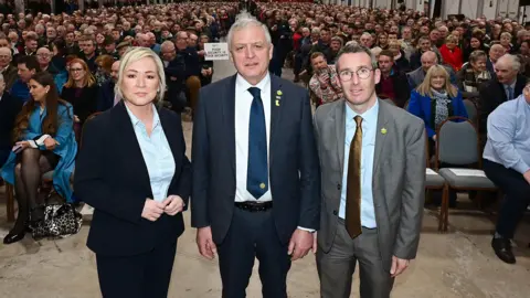 Pacemaker First Minister Michelle O'Neill, dressed in a navy trouser suit and blue shirt, on the left, with Agriculture Minister Andrew Muir, dressed in grey suit and brown tie, on the right. In the middle is William Irvine in a navy suit and navy tie. He has grey hair. The exhibition is full of people sitting on chairs.