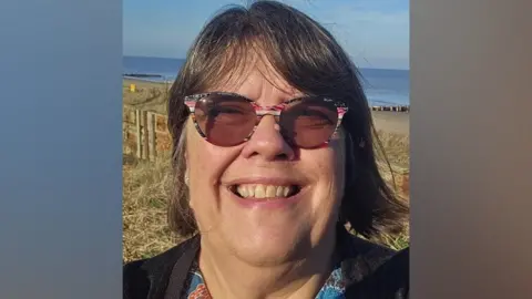 Julie Brociek-Coulton Julie Brociek-Coulton smiling and looking into the camera. She has short dark hair, and is wearing round tinted glasses with pink, black, and white patterned frames. In the background, there is grass and the sea.