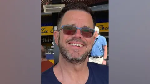 Wiltshire Police Jonathan Neilan smiling while sat outside under shade on a sunny day outside a restaurant. He is wearing a navy top and sunglasses.