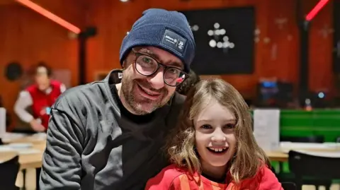 Supplied David Evans and his daughter Nina. David is wearing a grey jumper and Nina a pink jumper. They are sitting next to each other in what appears to be a cafe or restaurant. 
