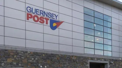 BBC Exterior of the Guernsey Post headquarters. The building has silver cladding and a set of 21 windows clustered together. A company logo is on the cladding. The lower half of the building is a stone wall.