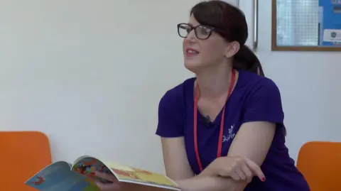 Mariam Badavi, wearing black framed glasses and a purple t-shirt, is sitting on an orange chair in an assembly room with her book in her hand as she looks out to the children who are asking her questions.