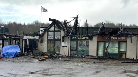 The units have been gutted by fire. The roof is gone and there are pieces of fire-damaged debris on the ground. On the left hand side is the car, which has been covered with a sheet of blue tarpaulin.