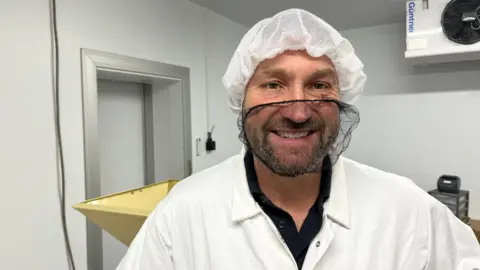 David Wheatley has a dark beard and moustache covered in a black hair net, he has a white hair net on his head and is wearing a white boiler suit with black shirt beneath, he is standing in the milling room, all white washed walls, with a hopper and machine behind him.