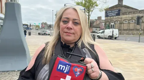 Margaret Oakes wearing black and holding a red bleed kit. She has long blonde hair and is standing on a paved street in Lowestoft town centre.
