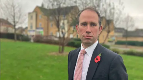 Matthew Barber is pictured on the Great Western Park estate in Didcot. He is standing in front of grass and trees and a small parade of shops can be seen in the background. He is wearing a grey suit, a salmon-coloured tie and a red poppy. 