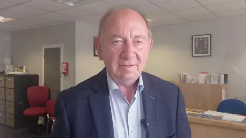 William Irwin is wearing a navy, checked suit jacket over a pale coloured shirt. He has grey hair and is standing in an office. 