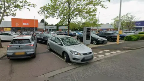 Google A B&Q site with its white and orange logo on the left of the photo is on a retail park with other businesses in the background. A car park is in the foreground, with two cars particularly prominent at a junction at the bottom of the image.