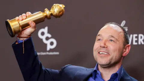 Reuters Stephen Graham smiles as he holds a Golden Globe award in the air, he wears a purple shirt under a blue blazer
