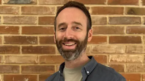 Peter Jenkins A man with short black hair and a beard, wearing a dark blue jacket and grey/green shirt, smiles as he stands in front of a brick wall.