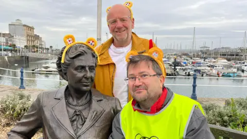 BBC Radio Devon presenter Michael Chequer and Matt Newbury from the Agatha Christie Festival are next to the Agatha Christie bench statue on Torquay harbour. The men and statue have Pudsey Bear ear Alice bands on.