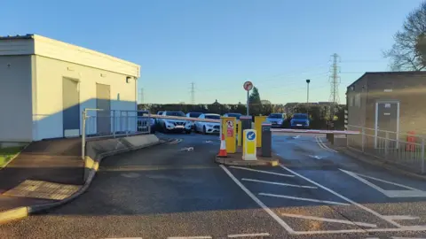 Cars parked in a car park in front of a hedge and behind an entrance with barriers across the entrance and exit lanes. A small brick building is on the right of the entrance with a white building on the left.