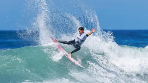A person on a pink surfboard is carving sharply across a wave, sending a large spray of water into the air. The ocean stretches into the background under a clear blue sky, with the surfer positioned mid‑turn on a breaking wave.