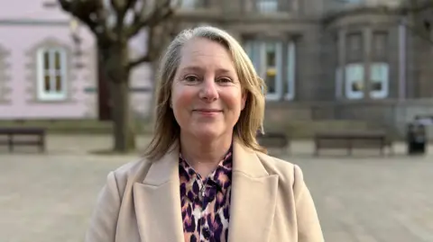 Sarah Hamon standing in Jersey’s Royal Square. She has long blonde hair and is wearing a leopard-print outfit underneath a camel-coloured coat. She is smiling directly at the camera.