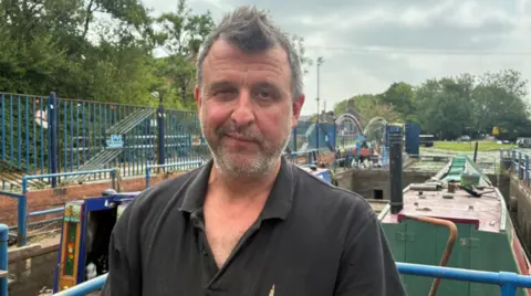A man with short brown hair, wearing an open-neck collared black shirt, looks at the camera. You can see canal boats in the background.