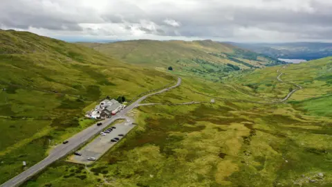 Terry Abraham Aerial image of the car park opposite the Kirkstone Pass Inn and holiday lets. The car park is rectangular shaped and has about 10 vehicles parked there. The Kirkstone Pass itself runs through green hills with a lake in the distance.