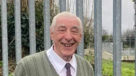 Luke Deal/BBC Mr Read smiles standing in front of a large metal gate. He wears a knitted green jumper with a white shirt underneath and a purple tie. He has grey hair. 