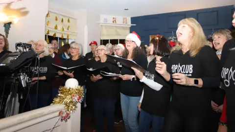 Barry Burns Photography A choir is singing from song sheets in a room with a blue wall. There is tinsel wrapped around a wooden post with santa decorations.