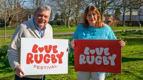 Rugby Borough Council Councillors Neil Sandison and Maggie O'Rourke stand in a park holding "Love Rugby" boards. It's a sunny day and there are trees and flowers around them.