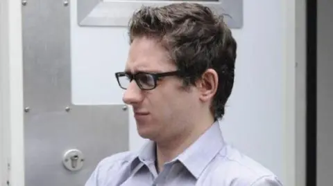 Spindrift A man with dark hair and glasses, wearing a light-coloured shirt, in a close-up shot. He is walking past a heavy security door