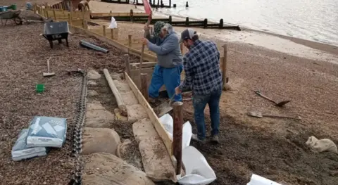 Two men trying to install some kind of barrier in the beach to halt the impact of coastal erosion. They are holding spades, there is a wheelbarrow, and bags of some type of grit. The sea is also in shot. 