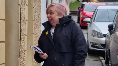 Lisa Pritchard A woman looking to the left of the image. She has white hair which has been dyed a light pink. She is door knocking and has leaflets in her right hand. She wears a dark blue coat and there are cars parked behind her on the street.