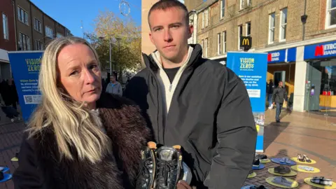A blonde woman in a fur coat holds a black pair of shoes. Beside her is her son Mani in a black coat with short hair. There are shoes on the ground behind them, and in the background High Street shops and people walking.