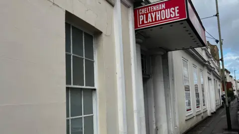 The side of a theatre building. The walls are painted a cream colour. There is a red sign above the door saying Cheltenham Playhouse.