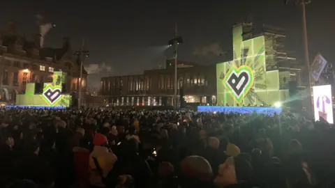 Thousands of people wearing woolly hats standing in City Park in Bradford looking at two illuminated stages lit up with a big letter B in black surrounded by a yellow, green and pink background.   