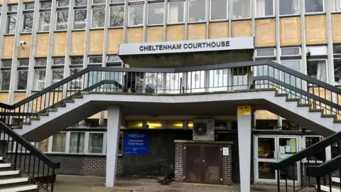The entrance of Cheltenham Magistrates' Court. It is a 1960's building and the entrance in on the first floor, accessed by two flights of stairs on either side. Above the entrance doors is a sign that reads "Cheltenham Courthouse".