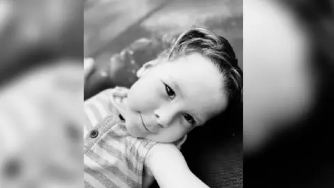 Sian Channon A black and white image of a young boy looking at the camera wearing a stripey sleeveless baby grow.