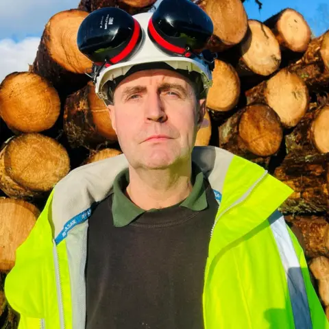 Duncan Cassie looking at the camera, he has a bright yellow jacket on, and ear protectors are on his white helmet. He is standing in from of chopped down timber.
