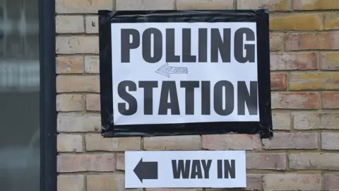 A generic sign stuck on a brick wall that reads polling station. Another sign below it details the way inside the polling station to the left. 