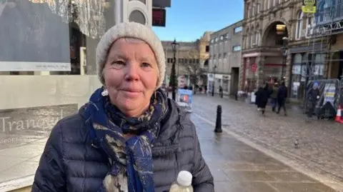 Gemma Dillon/BBC A woman wearing a black padded jacket and navy blue scarf with cream hat stands on a cobbled street with shops behind her.