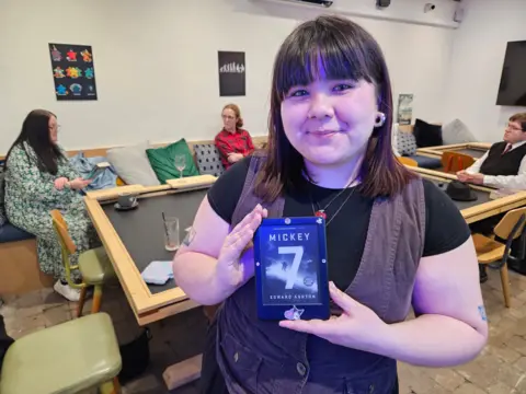 A woman with dark brown hair holds an e-reader displaying the book Mickey 7 by Edward Ashton.