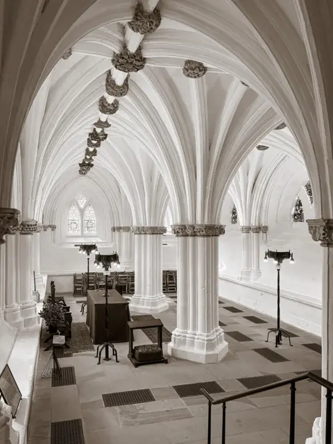 David Smith An ornate stone hall with soaring Gothic vaulted ceilings and carved columns stretches into the distance. Decorative stone bosses line the ceiling, while tables, chairs and tall lamps sit beneath arched windows that let light into the historic interior.