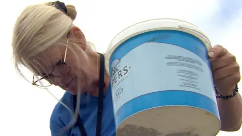 A woman is looking down and feeding animals from a feed tub. She has blonde hair and glasses and is wearing a blue t shirt
