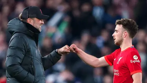 PA Media Jurgen Klopp, wearing a black Liverpool FC branded bubble coat and a black cap, smiles as he fist bumps Diogo Jota, who is wearing his red Liverpool kit, during a match
